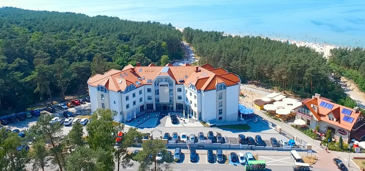 Widok na hotel White z lotu ptaka, otoczony lasem i blisko plaży. Na pierwszym planie parking z samochodami, a w tle morze i plaża. Idealne miejsce na wakacje.