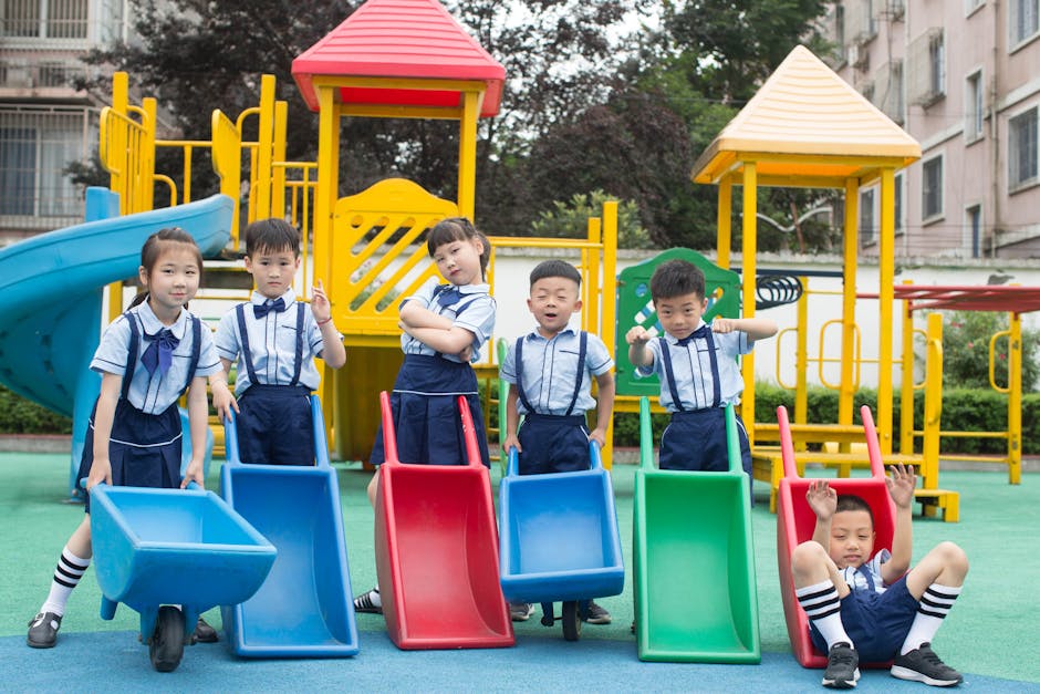 Grupa sześciu dzieci w szkolnych mundurkach bawi się na placu zabaw, trzymając kolorowe zjeżdżalnie. W tle widoczne są huśtawki i zjeżdżalnie. Dzieci pozują w radosnych pozach, co podkreśla atmosferę zabawy i przyjaźni.