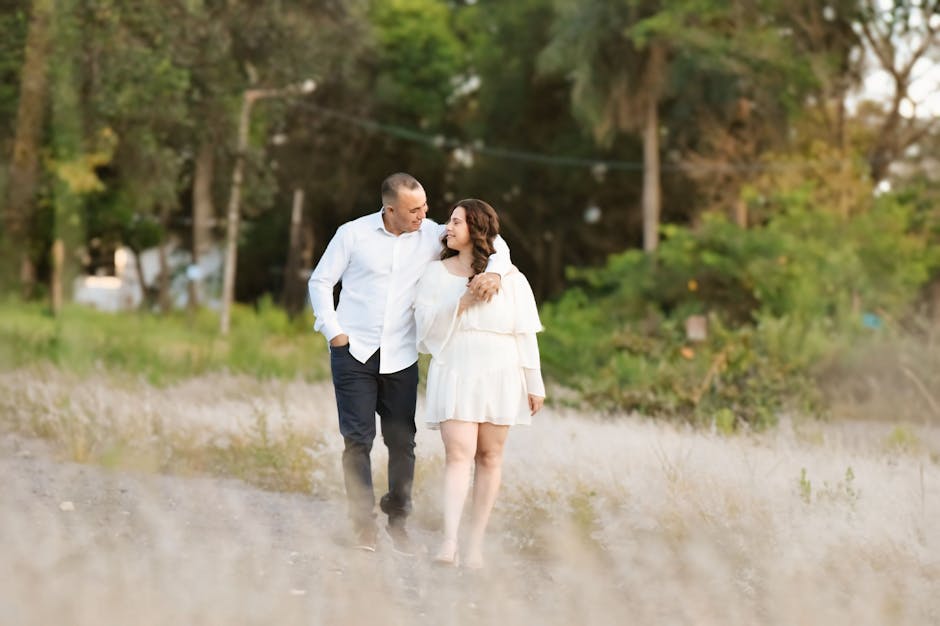Para spacerująca po polu, uśmiechająca się do siebie. Mężczyzna w białej koszuli i ciemnych spodniach, kobieta w białej sukience. Tło z zielonymi drzewami i delikatnym oświetleniem.