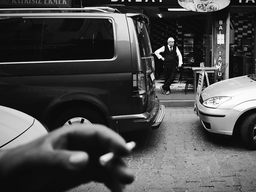 Mężczyzna w eleganckim stroju stoi na zewnątrz restauracji, otoczony samochodami. W tle widoczny jest szyld lokalu. Scena utrzymana w czarno-białej kolorystyce, z wyraźnym akcentem na ruch uliczny i codzienne życie w mieście.