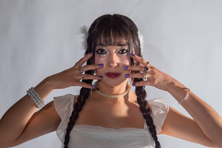 Młoda kobieta z warkoczami, w białej sukience, prezentująca zdobienia na dłoniach i twarzy, z błyszczącym makijażem oraz biżuterią. Tło w jasnym kolorze.