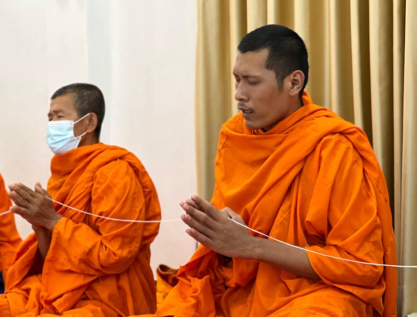 Mnisi w pomarańczowych szatach modlą się w skupieniu, siedząc w tradycyjnej pozycji. W tle widoczne są zasłony oraz elementy wnętrza, co podkreśla atmosferę medytacji i duchowości.
