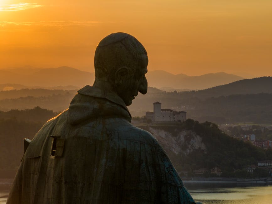 Pomnik postaci z profilu, z zachodzącym słońcem w tle, nad malowniczym krajobrazem górskim i zamkiem.
