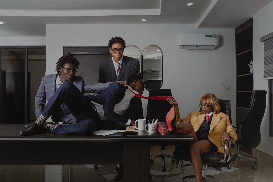 Czterech biznesmenów w garniturach w biurze, z jednym mężczyzną leżącym na stole, a kobieta w żółtym garniturze trzymająca wysokie obcasy, podczas gdy pozostali mężczyźni obserwują sytuację.